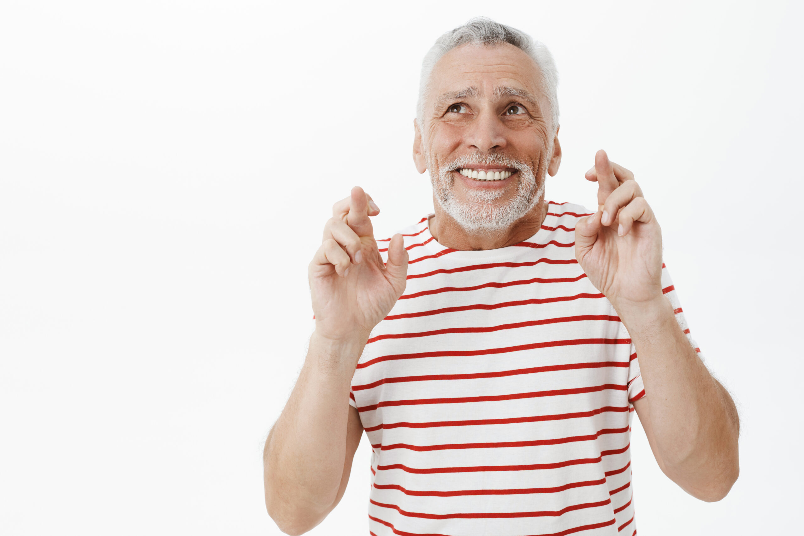 Optimistic happy and charismatic old male pensioner in striped t-shirt raising hands crossing fingers for good luck looking at upper left corner with hopeful smile making wish over white wall. Copy space